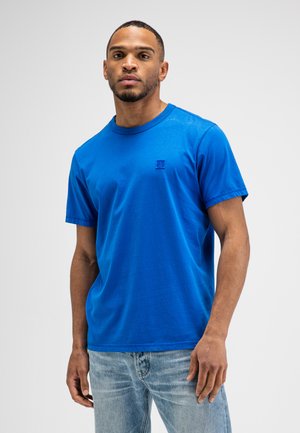 Man draagt een effen blauw T-shirt met korte mouwen en lichtblauwe spijkerbroek, staand tegen een witte achtergrond.
