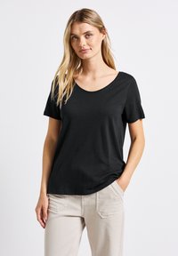 Černé tričko s krátkým rukávem a kulatým výstřihem, vyrobené z měkké látky, v kombinaci se světle béžovými kalhotami. Minimalistický design bez vzorů.