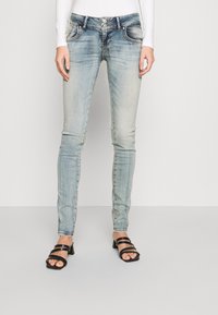 Lightly faded blue skinny jeans with two front pockets, dual-button closure, and a slim fit. Paired with black strappy heeled sandals.