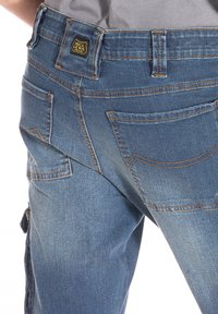 Pantalons cargo en denim bleu avec des coutures visibles, plusieurs poches et une étiquette de marque sur la taille à l'arrière. Caractéristiques d'une coupe relaxée.