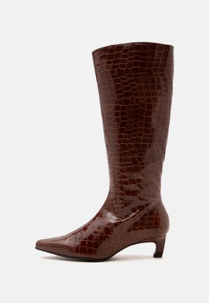 Botte haute marron foncé avec texture crocodile, bout pointu et petit talon kitten sur un fond clair uni.
