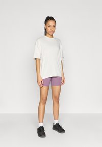 Ανοιχτό γκρι φαρδιά t-shirt με μικρό λογότυπο στο στήθος, σε συνδυασμό με μοβ ποδηλατικά σορτς και μαύρα αθλητικά παπούτσια με λευκές κάλτσες.