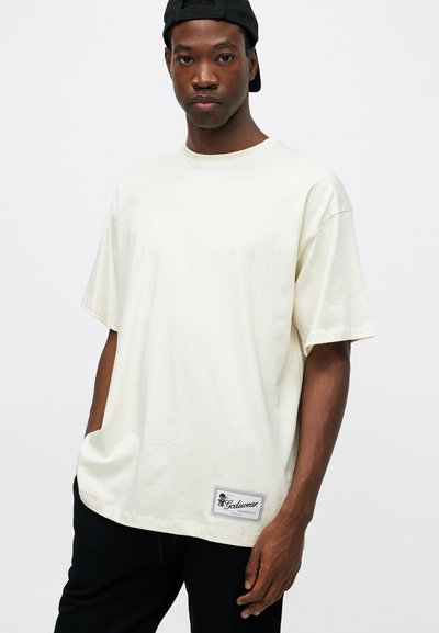 Cream-colored oversized t-shirt made of soft cotton, featuring a label at the bottom left, with short sleeves and a round neckline.