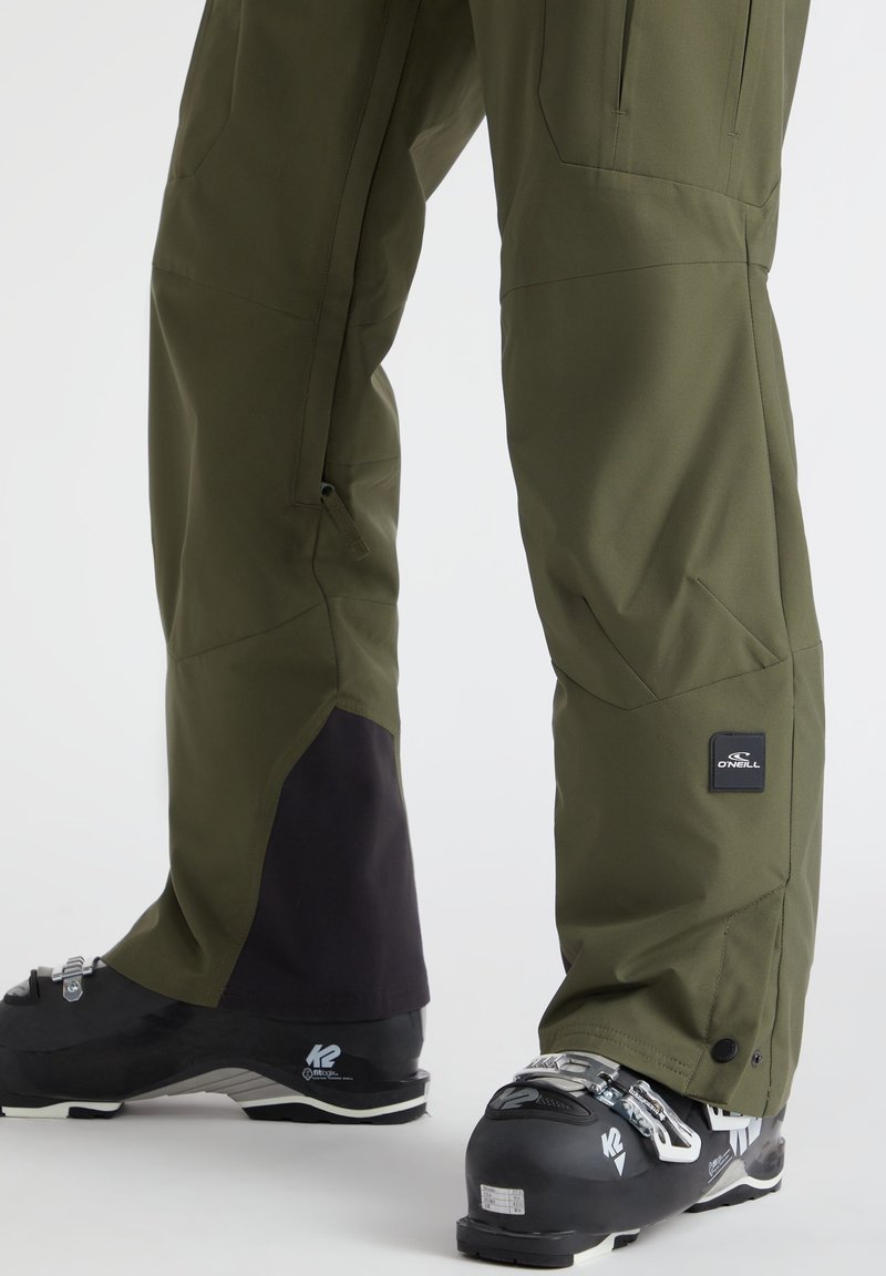 Grüne Outdoor-Hosen mit tailliertem Design, schwarzen Abschlüssen, Seitentaschen und einem Logo-Patch. Getragen mit schwarzen Skischuhen.