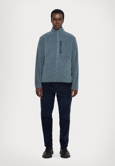 Fleecejacke in Hellblau mit hohem Kragen, Reißverschluss, Fronttasche und kontrastierenden schwarzen Details. Kombination mit dunklen Hosen und Schuhen.