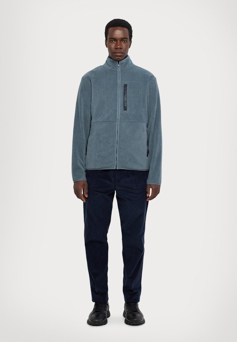 Veste en polaire bleu clair avec un col haut, fermeture éclair, poche frontale et détails noirs contrastants. Associée à un pantalon et des chaussures foncés.