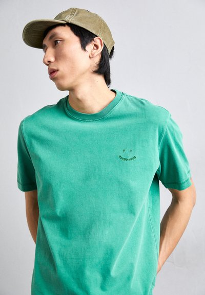 Πράσινο βαμβακερό μπλουζάκι με στρογγυλή λαιμόκοψη και κοντά μανίκια. Διαθέτει μια μικρή γραφική απεικόνιση χαρούμενου προσωπου σε σκούρο πράσινο χρώμα στο στήθος. Ρυθμισμένο για χαλαρή εφαρμογή.