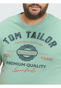 Mintgrünes T-Shirt aus Baumwolle mit rundem Ausschnitt und schwarz-rotem Siebdruck-Logo und Text auf der Vorderseite.