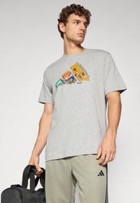 Grå bomull T-skjorte med et fargerikt grafisk design med bygninger og en figur. Modellen holder en svart treningsbag.