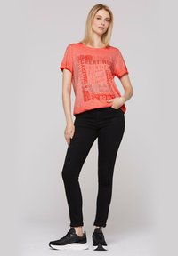 Camiseta de manga corta color coral con texto gráfico, combinada con jeans negros ajustados y zapatillas negras. Estilo de outfit casual y cómodo.