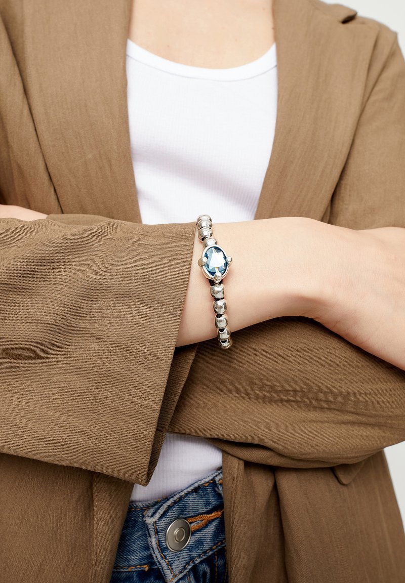 Pulsera de plata con cuentas redondas y una gran piedra preciosa azul en el centro, sobre un fondo de tela marrón texturizada.