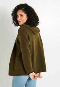 Cardigan à capuche vert olive avec une coupe ample, en tissu texturé, deux poches avant et des poignets côtelés. Porté sur un haut noir et un jean clair.