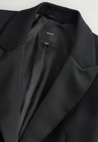 Zwarte blazer met een gladde satijnen voering, scherpe pieklapels en een rechthoekig merketiket met "NEXT" binnenin de kraag.