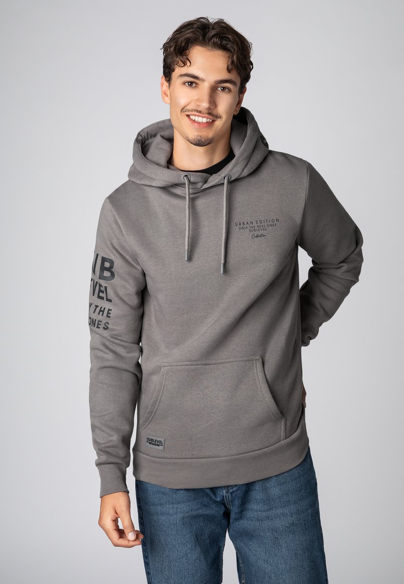 Grijze hoodie van zacht materiaal. Heeft een voorzak, verstelbare koordjes en bedrukte tekst op de borst en mouw.