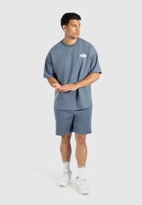 Blå bomulds T-shirt og matchende shorts med en afslappet pasform og minimalistisk design, prydet med et lille grafisk logo på brystet. Hvide sneakers.