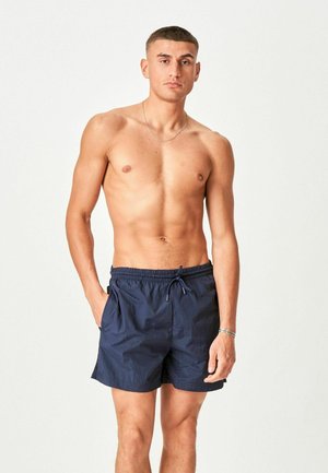 Mladý muž bez trička, oblečený v tmavomodrých plavkových šortkách so sťahovacou šnúrkou, stojaci s jednou rukou v vrecku na jednoduchom svetlom pozadí.