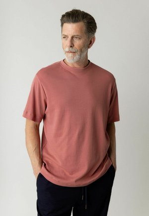 Muž so sivou bradou, oblečený v ružovej krátkosleevej košeli a tmavých nohaviciach, hľadí doľava s rukami v nohaviciach na jednofarebnom pozadí.