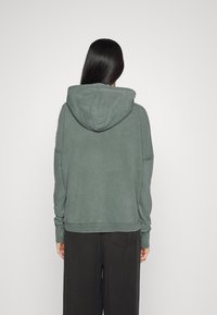 Donna con lunghi capelli scuri, che indossa un felpa con cappuccio verde e pantaloni neri, in piedi di fronte a uno sfondo chiaro e semplice.