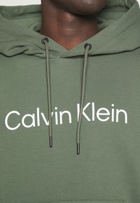 Felpa verde oliva realizzata in misto cotone, con tasca frontale a marsupio, corde regolabili, e logo grafico bianco "Calvin Klein".
