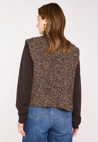 Bruin gestructureerd vest met een gebreide achterkant en lange mouwen in een donkerdere tint, gecombineerd met een blauwe spijkerbroek. Heeft een rughalslijn met een ronde uitsnijding.