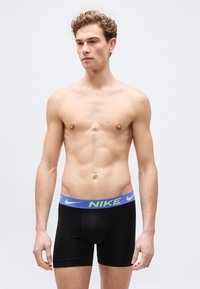 Νέος άνδρας με ανοιχτόχρωμο δέρμα και σγουρά μαλλιά, φοράει μαύρα μποξεράκια Nike με μπλε και πράσινη μέση, στέκεται μπροστά σε μια απλή ανοιχτόχρωμη επιφάνεια.