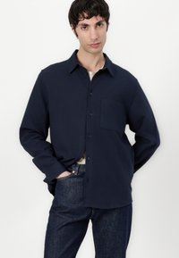 Chemise boutonnée bleu marine en tissu doux, avec un col, une poche poitrine unique et des manches longues, assortie à un jean en denim foncé.
