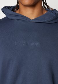 Námornícky modrá mikina s kapucňou so štruktúrovaným povrchom, na prednej strane s embossovaným logom "Calvin Klein", vyrobená z mäkkej látky.