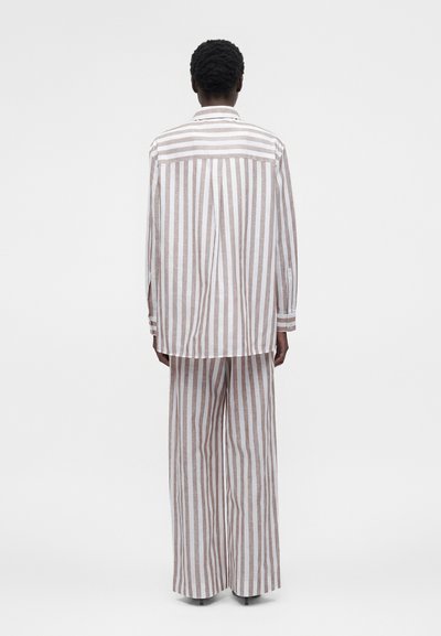 Άτομο από πίσω με φαρδιά, μπεζ και λευκή ριγέ πουκαμίσα με μακριά μανίκια και ταιριαστό παντελόνι με φαρδιά πόδια σε απλό φόντο.