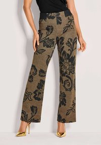 Frau trägt braune weit geschnittene Hosen mit schwarzen Blumenmustern und goldenen High-Heels, steht vor einem hellen Hintergrund.