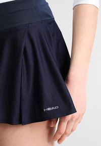 Falda deportiva azul marino hecha de tela de malla transpirable, con una cinturilla ancha y el logo "HEAD" en gris en el dobladillo.
