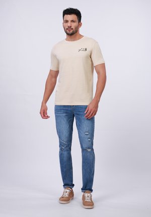 Beige Baumwoll-T-Shirt mit kurzen Ärmeln und einem kleinen Druckdesign auf der Brust, kombiniert mit blauen, abgeranzten Jeans und beigen Sneakers.