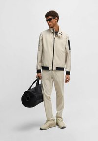 Giacca impermeabile beige con dettagli neri, chiusura a zip e colletto, abbinata a pantaloni crema e sneaker beige, con una borsa duffel nera.