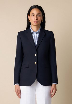 Frau mit langen dunklen Haaren trägt ein marineblaues Blazer mit goldenen Knöpfen über einem blau-weiß gestreiften Hemd, kombiniert mit weißen Hosen.