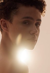 Skulderlangt krøllete hår, høydepunkt på kinnbenet, myk hudtekstur, belyst bakfra med et sterkt lys som skaper en halo-effekt rundt figuren.