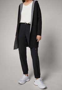 Svart lång cardigan med grå huva, ihop med svarta tapered byxor och vita sneakers. Tyget verkar mjukt, med fickor på byxorna.