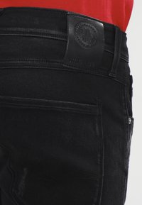 Svarta denimjeans med en texturerad yta, en läderetikett vid midjan och subtila sömdetaljer. Flera synliga fickor.