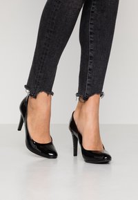 Zapatos de tacón alto de charol negro con punta redonda y parte trasera abierta, con un tacón texturizado. Llevados con unos jeans de mezclilla negra deshilachados.