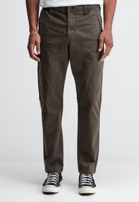 Bruine chinos van een zachte, geweven stof, met een knoopsluiting, elastische tailleband en zijzakken. Gecombineerd met zwarte canvas sneakers.