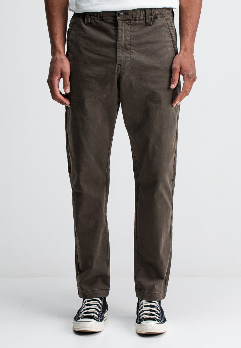 Bruine chinos van een zachte, geweven stof, met een knoopsluiting, elastische tailleband en zijzakken. Gecombineerd met zwarte canvas sneakers.