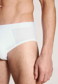 Witte katoenen slips met een geribbelde textuur, voorzien van een zachte elastische tailleband. Close-up weergave die het zijprofiel en een deel van een hand toont.