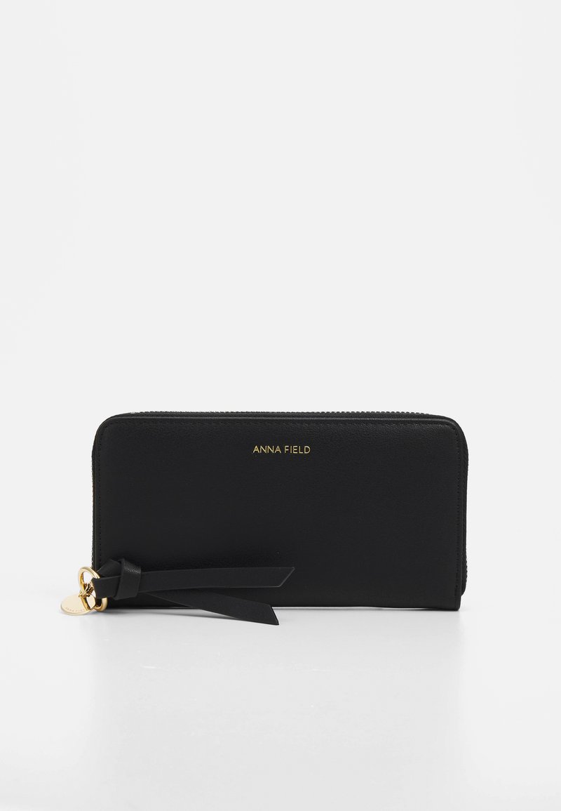 Portefeuille noir zippé en matériau synthétique lisse, avec un logo doré et un gland décoratif agrémenté d'un charme en métal.