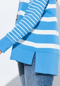 Felpa a righe blu e bianche con polsini e orlo a costine, dotata di spacchi laterali per una maggiore libertà di movimento, abbinata a jeans azzurri.