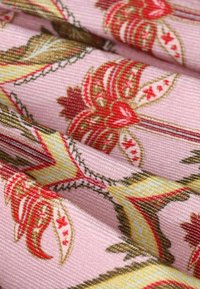 Tissu rose avec des motifs floraux et géométriques rouges, jaunes et marron complexes, présenté en gros plan avec des plis mettant en valeur la texture et le design.