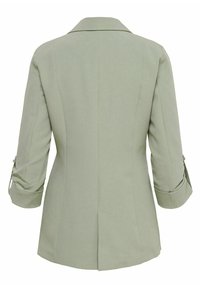 Blazer vert clair pour femme avec manches trois quarts et fente arrière, vu de dos sur un fond uni.