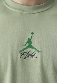 Silhouette verte brodée d’un joueur de basketball sautant avec un ballon au-dessus de la tête et texte « Flight » en dessous sur un tissu vert clair.