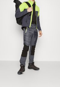 Veste de sport bleu marine et vert néon avec fermeture éclair à l'avant ; pantalon isolé gris avec accents noirs ; chaussures noires robustes ; sac à dos noir.