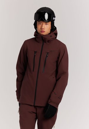 Man draagt een bruine skipak en broek, zwarte skihandschoenen en een zwarte skihelm met bril, staande tegen een effen achtergrond.