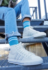 Bílé vysoké tenisky s modrými pruhy, texturovaná kůže a bílé gumové podrážky. Spárováno s modrými pruhovanými ponožkami, sedící na kovových schodech.