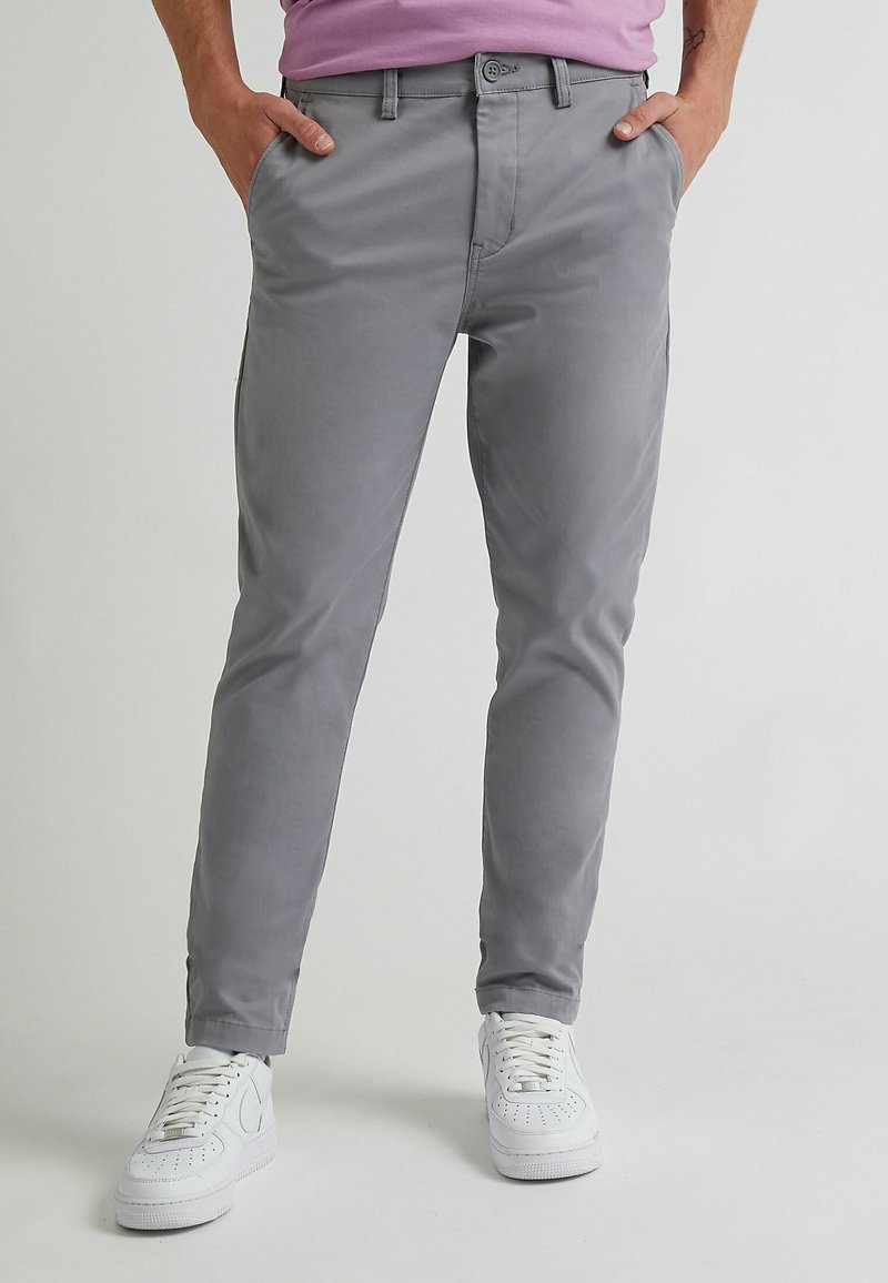 Personne portant un pantalon slim gris clair et des baskets blanches, debout les mains dans les poches devant un fond uni.