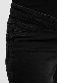 Jeans de denim negro con una vista en primer plano, que presentan una superficie texturizada, una cinturilla desigual y un pequeño detalle de bolsillo delantero.